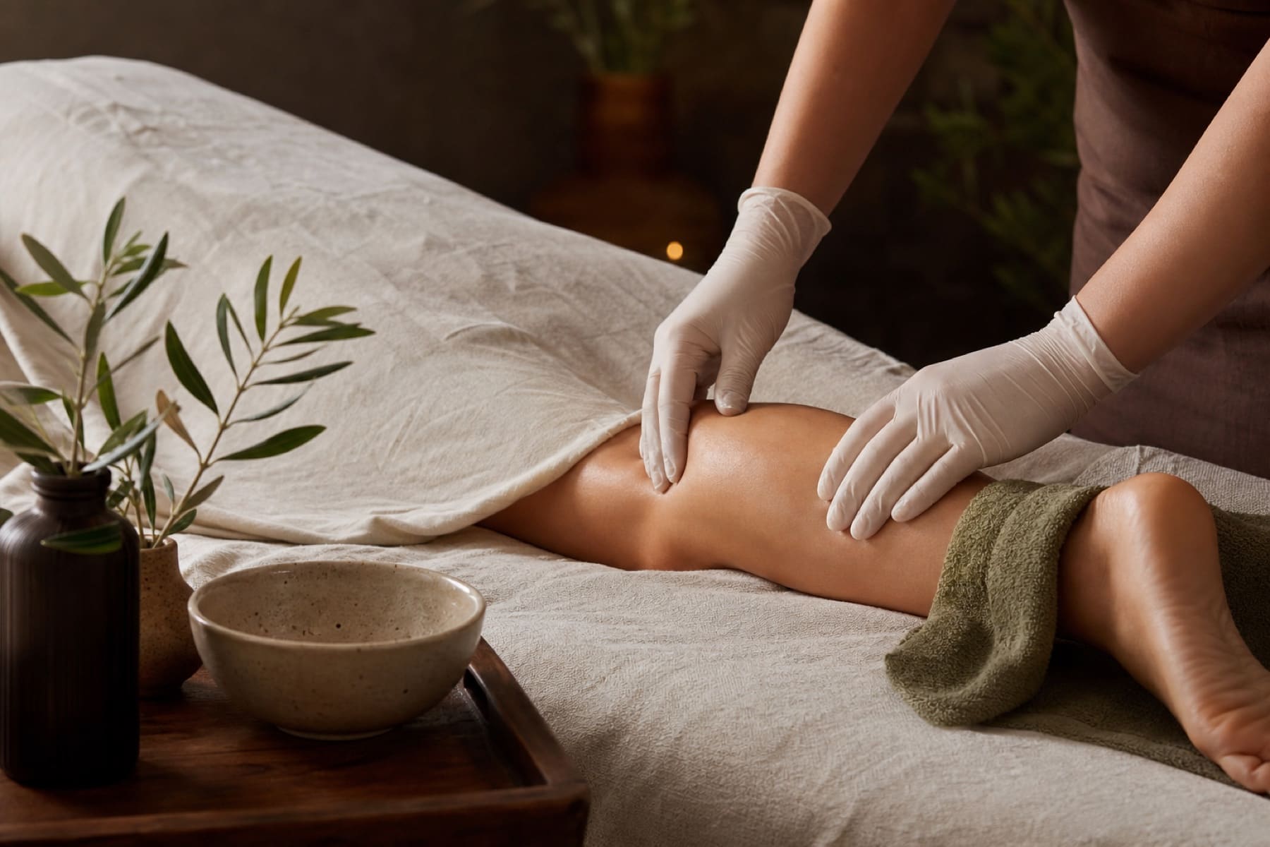 Gentle lymphatic massage setup with gloved hands, cream linen, and soft olive spa accents.